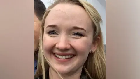 Andrea Brady Un primer plano de Jessica Brady, sonriendo ampliamente y mirando directamente a la cámara, con cabello largo y rubio, ojos marrones y un arete visible en su oreja izquierda.