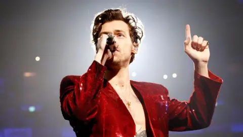 JMInternational/Getty Images Harry Styles canta frente a un micrófono mientras actúa en los Brit Awards 2023. Lleva una chaqueta roja de lentejuelas y señala con el dedo el techo.
